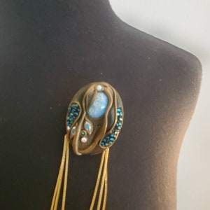 May include: An oval-shaped brooch with a bronze-colored frame, adorned with a central light blue stone and smaller blue and clear crystals. Gold-colored chains hang from the bottom, adding a decorative element.