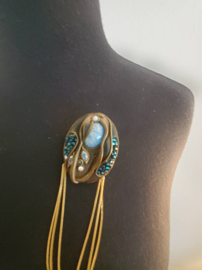 May include: An oval-shaped brooch with a bronze-colored frame, adorned with a central light blue stone and smaller blue and clear crystals. Gold-colored chains hang from the bottom, adding a decorative element.
