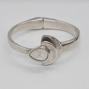 May include: A silver-toned hinged bangle bracelet. The bracelet features a pear-shaped, iridescent stone set in a crescent-shaped design. The bracelet has a smooth, polished finish and a clasp closure. The brand name is engraved on the bracelet.