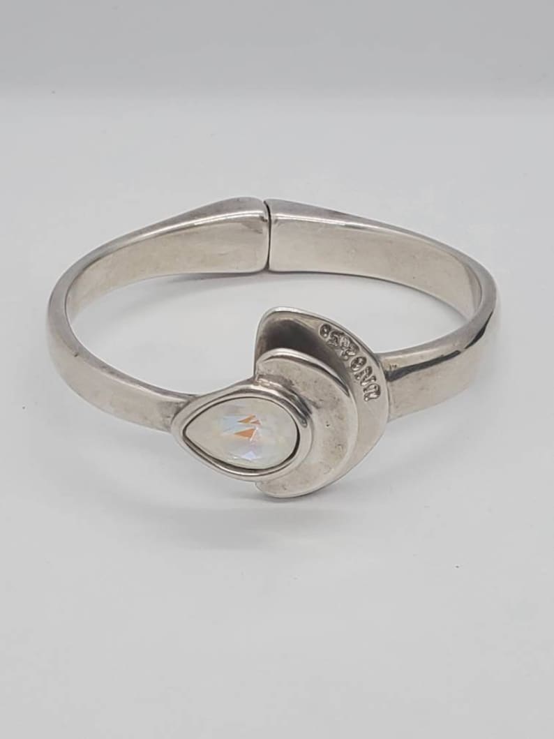 May include: A silver-toned hinged bangle bracelet. The bracelet features a pear-shaped, iridescent stone set in a crescent-shaped design. The bracelet has a smooth, polished finish and a clasp closure. The brand name is engraved on the bracelet.