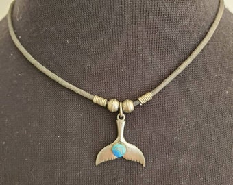 May include: A silver-toned necklace featuring a whale tail pendant with a blue stone inlay. The pendant hangs from a gray cord necklace with silver beads. The necklace is displayed on a black background.