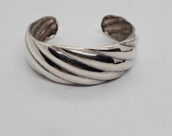 May include: A silver-colored cuff bracelet with a ridged, textured design. The open-ended bracelet has a polished, reflective surface and a curved shape, suitable for wrist wear. The jewelry piece is set against a white background.