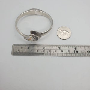 May include: A silver-toned hinged bangle bracelet with an abstract design. The bracelet features a central decorative element with a light-colored, possibly mother-of-pearl, inlay. A ruler shows the bracelet is approximately 2.5 inches in diameter.