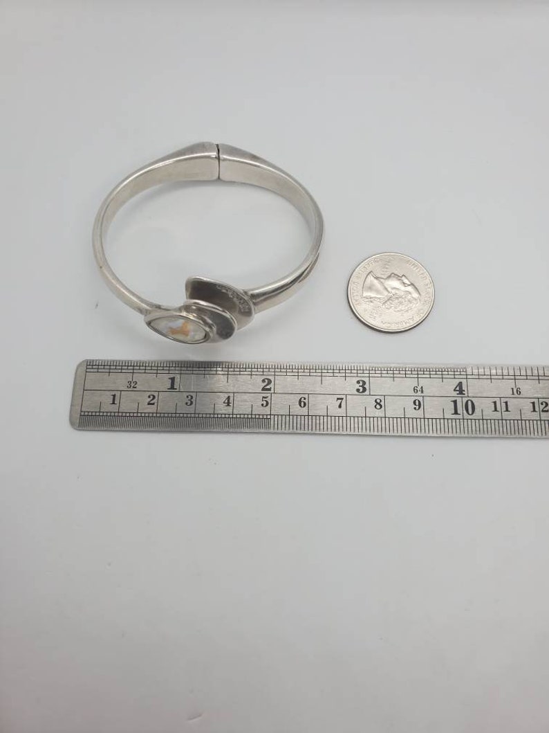 May include: A silver-toned hinged bangle bracelet with an abstract design. The bracelet features a central decorative element with a light-colored, possibly mother-of-pearl, inlay. A ruler shows the bracelet is approximately 2.5 inches in diameter.
