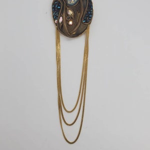May include: An oval-shaped, bronze-toned brooch with a central design featuring iridescent and blue accents. Three strands of gold-colored chain hang from the brooch, creating a layered effect. The brooch has a vintage aesthetic.