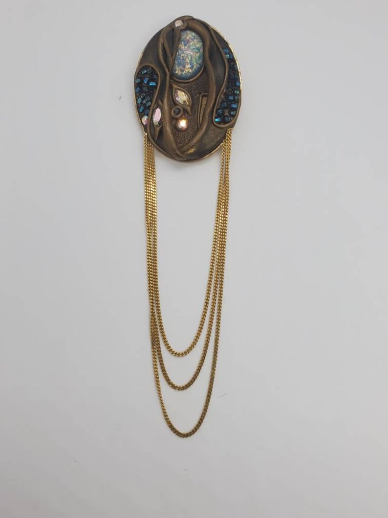 May include: An oval-shaped, bronze-toned brooch with a central design featuring iridescent and blue accents. Three strands of gold-colored chain hang from the brooch, creating a layered effect. The brooch has a vintage aesthetic.