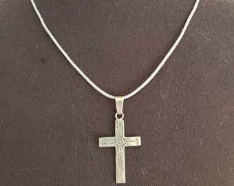 May include: A silver-colored cross pendant necklace. The cross has etched details and hangs from a thin, white cord. The pendant is centered against a dark background.