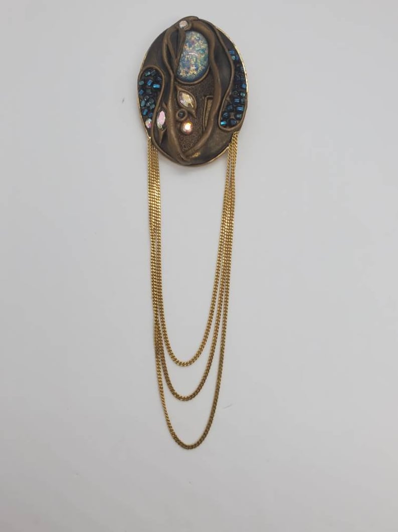 May include: An oval-shaped, bronze-toned brooch with a central design featuring iridescent and blue accents. Three strands of gold-colored chain hang from the brooch, creating a layered effect. The brooch has a vintage aesthetic.