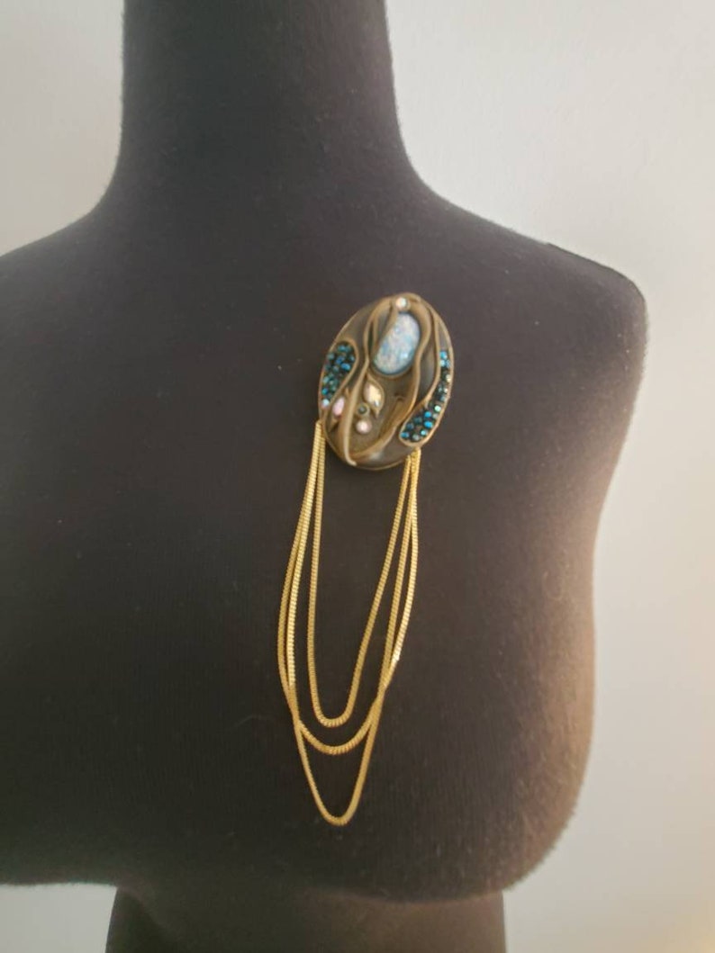 May include: An oval-shaped brooch with a bronze-colored base, adorned with blue and pink gemstones and a central light blue stone. Three strands of gold-colored chain hang from the brooch, creating a layered effect. The brooch is displayed on a black mannequin.