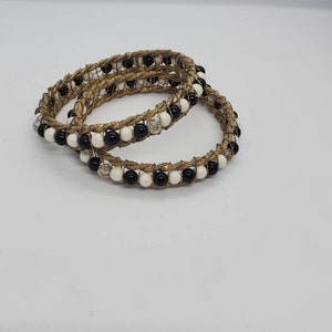 May include: Two gold-toned bracelets with a woven design. Each bracelet is adorned with alternating black and white beads, interspersed with clear, faceted beads. The bracelets are displayed on a white surface.