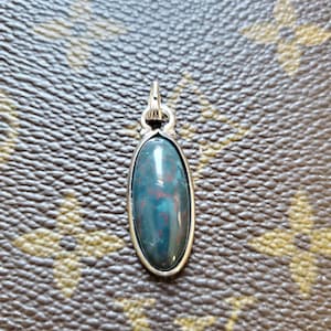 May include: Oval pendant featuring a polished, dark green gemstone with red inclusions, set in a silver-toned metal frame. The pendant has a loop at the top for a chain. The background is a brown, textured surface.