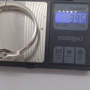 May include: A silver-colored bracelet is placed on a digital scale, displaying a weight of 39.5 grams. The scale is black with a blue digital display. The bracelet is curved and has a smooth, reflective surface.