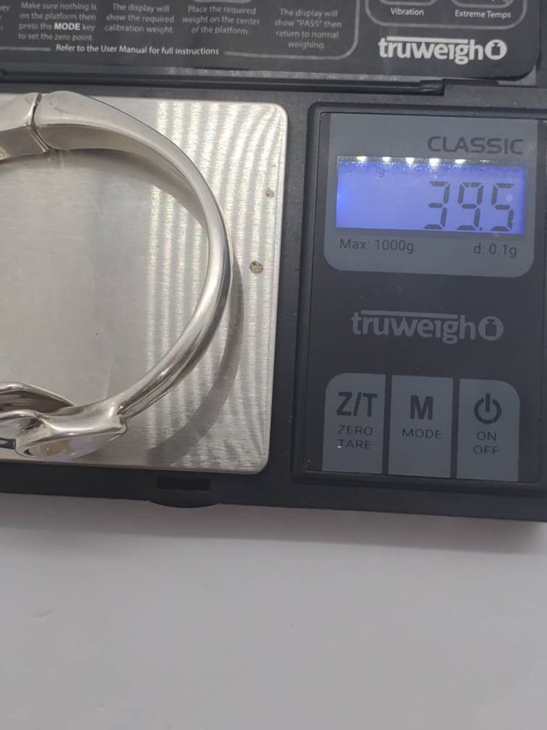 May include: A silver-colored bracelet is placed on a digital scale, displaying a weight of 39.5 grams. The scale is black with a blue digital display. The bracelet is curved and has a smooth, reflective surface.