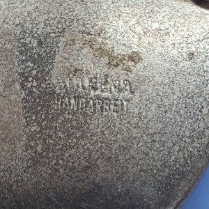 May include: Close-up of a vintage silver-toned brooch with the text "MARINA HANDARBEIT" embossed on the surface. The brooch has a textured, aged appearance, with a chain detail at the bottom. The brooch is heart-shaped.