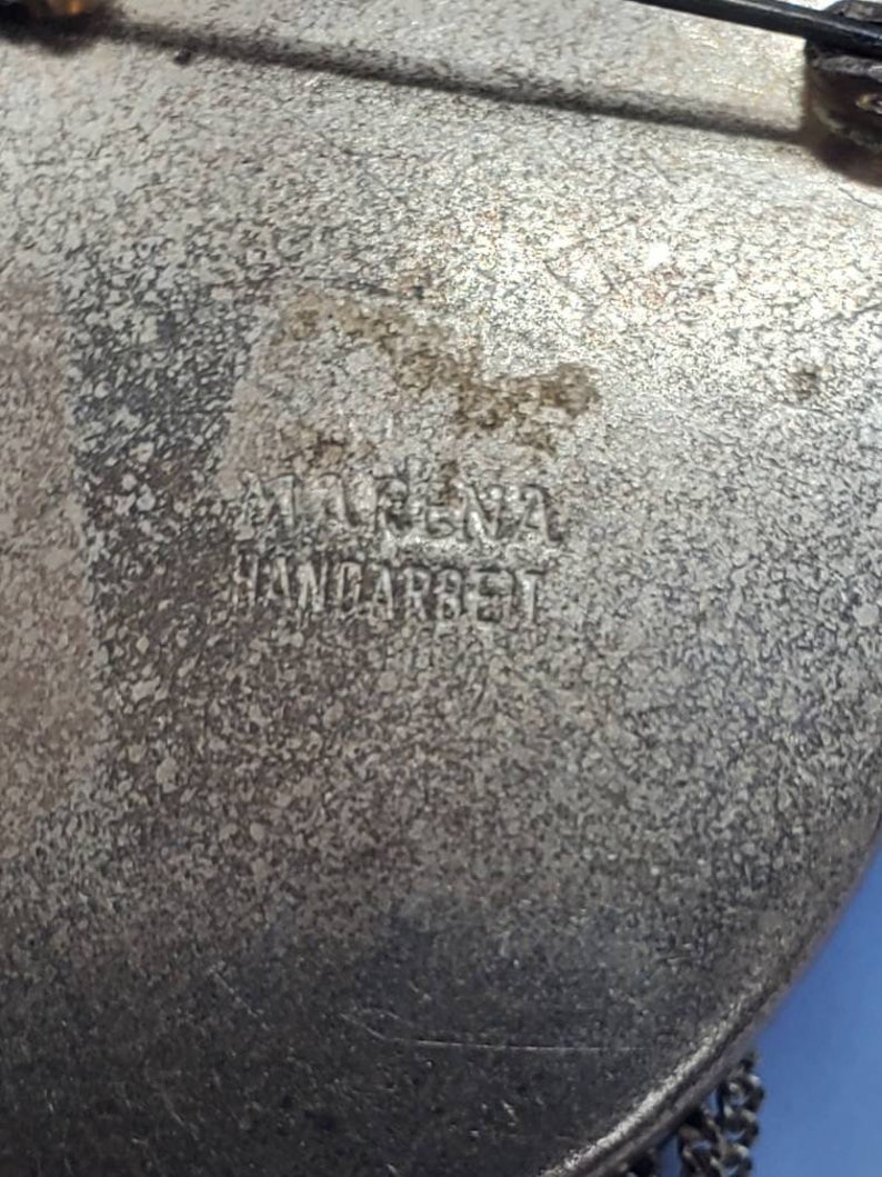 May include: Close-up of a vintage silver-toned brooch with the text "MARINA HANDARBEIT" embossed on the surface. The brooch has a textured, aged appearance, with a chain detail at the bottom. The brooch is heart-shaped.