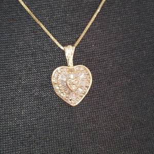 Puede incluir: Un collar dorado con un colgante en forma de coraz&oacute;n. El colgante est&aacute; adornado con peque&ntilde;as piedras transparentes y presenta un dise&ntilde;o en forma de coraz&oacute;n en el centro. La cadena es de tipo caja fina.