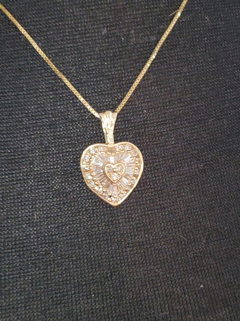 Puede incluir: Un collar dorado con un colgante en forma de coraz&oacute;n. El colgante est&aacute; adornado con peque&ntilde;as piedras transparentes y presenta un dise&ntilde;o en forma de coraz&oacute;n en el centro. La cadena es de tipo caja fina.