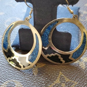 May include: A pair of gold-toned hoop earrings. Each earring features a circular design with blue, black, and silver-toned geometric inlays. The earrings are displayed on a black stand against a brown patterned background.