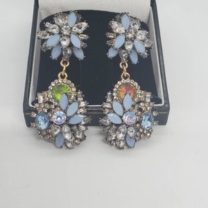 May include: A pair of ornate earrings with a floral design. Each earring features light blue petals, clear crystals, and iridescent gemstones. The earrings are displayed in a black jewelry box.