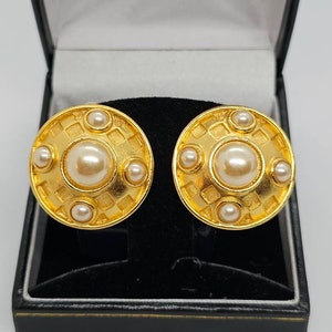 May include: A pair of gold-toned, round clip-on earrings with a central faux pearl surrounded by smaller pearls. The earrings have a textured, geometric design and are displayed in a black jewelry box.
