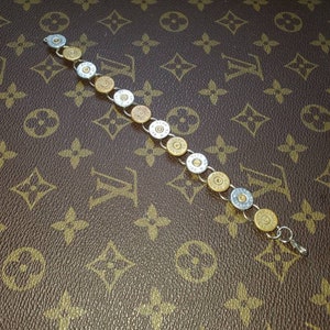 May include: A bracelet made of repurposed bullet casings. The bracelet alternates between silver and gold-toned casings, connected by a silver chain. The clasp is silver. The background is a brown leather-like surface with a repeating pattern.