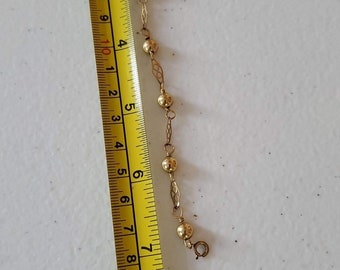 May include: A delicate gold-colored bracelet with small, round beads and decorative links. The bracelet is approximately 7 inches long, as measured by a yellow ruler. The jewelry is displayed on a white surface.