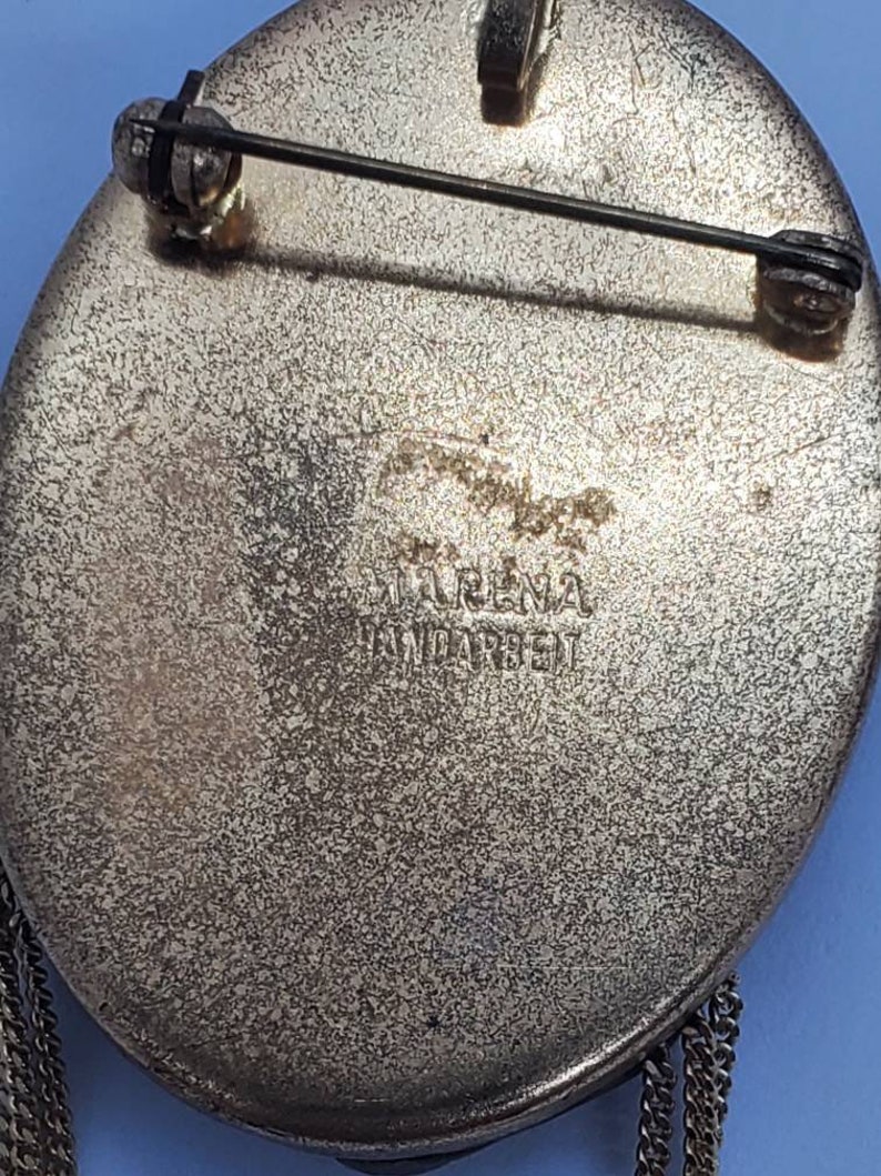 May include: Vintage oval brooch with a textured silver-tone finish. The back features a pin clasp and the inscription "IRENA HANDARBEIT". Decorative chain fringe hangs from the bottom of the brooch. A classic piece of jewelry.