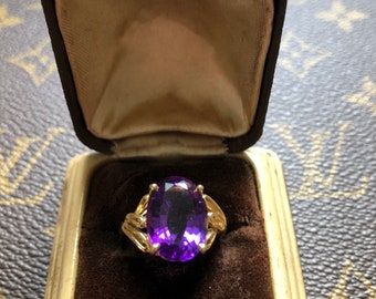 May include: A gold ring with a large, oval-cut purple gemstone, set in a decorative gold band. The ring is displayed in a vintage, open jewelry box with a brown velvet interior and a cream-colored exterior.