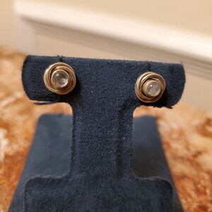 May include: A pair of stud earrings featuring a small, clear gemstone set in a gold-colored, spiral-wrapped setting. The earrings are displayed on a dark blue stand, showcasing their delicate design and craftsmanship.