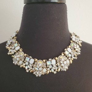 May include: A statement necklace with a gold-tone metal base, adorned with an array of iridescent and white gemstones. The necklace is displayed on a black mannequin, showcasing its intricate design and sparkling details. A glamorous accessory.