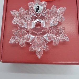 May include: A clear crystal snowflake ornament with intricate detailing. The ornament has a red ribbon for hanging and is on a red box. The ornament has the word "WATERFORD" and a seahorse logo.