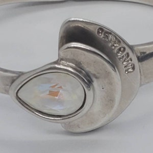 May include: Silver-toned hinged bracelet with a pear-shaped, iridescent crystal centerpiece. The bracelet features a unique design with a curved, layered element near the crystal. The brand name is visible on the bracelet.