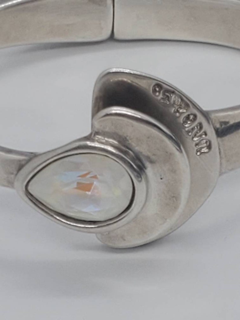 May include: Silver-toned hinged bracelet with a pear-shaped, iridescent crystal centerpiece. The bracelet features a unique design with a curved, layered element near the crystal. The brand name is visible on the bracelet.