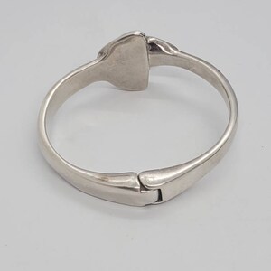 May include: A silver-toned hinged bangle bracelet. The bracelet has a rectangular center piece and a clasp closure. The metal has a polished finish, reflecting light. The design is simple and elegant, suitable for everyday wear.