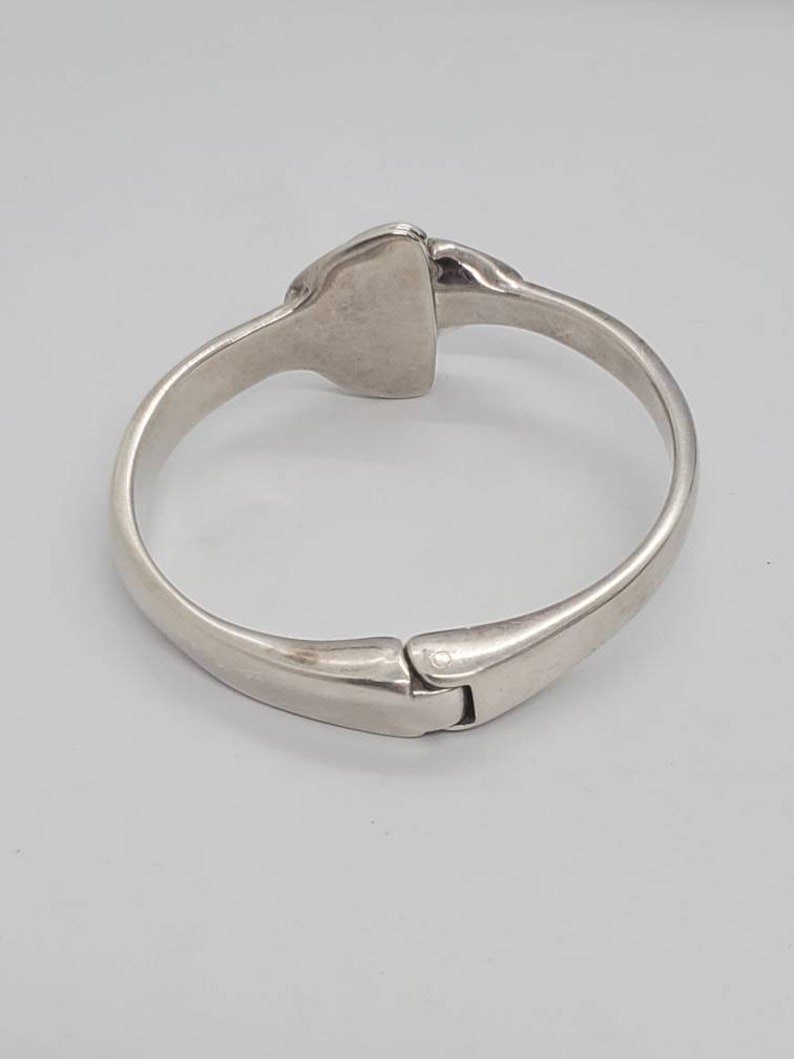 May include: A silver-toned hinged bangle bracelet. The bracelet has a rectangular center piece and a clasp closure. The metal has a polished finish, reflecting light. The design is simple and elegant, suitable for everyday wear.