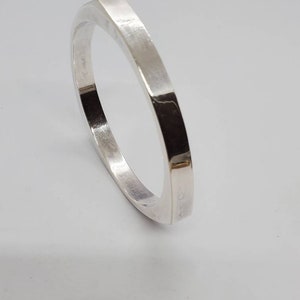 May include: A silver-colored, square-shaped bangle bracelet. The bracelet has a flat, brushed metal finish and a geometric design. The bracelet is approximately 2.5 inches in diameter.