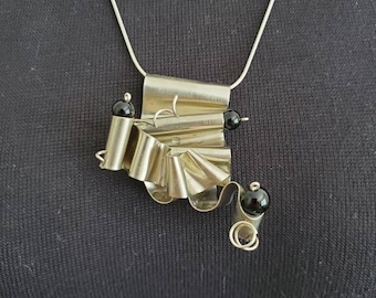 May include: Silver-toned metal necklace with a unique, abstract pendant. The pendant features a folded, ruffled design with two black beads and a delicate silver chain. The necklace is displayed against a dark background.