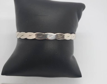May include: A silver-colored braided bracelet displayed on a black cushion. The bracelet has a woven design and appears to be made of metal. The bracelet is approximately 7 inches in length.