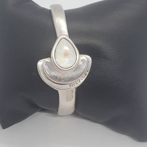 May include: A silver-toned cuff bracelet with a pear-shaped, iridescent crystal centerpiece. The crystal is set within a fan-shaped design. The bracelet is displayed on a black surface.