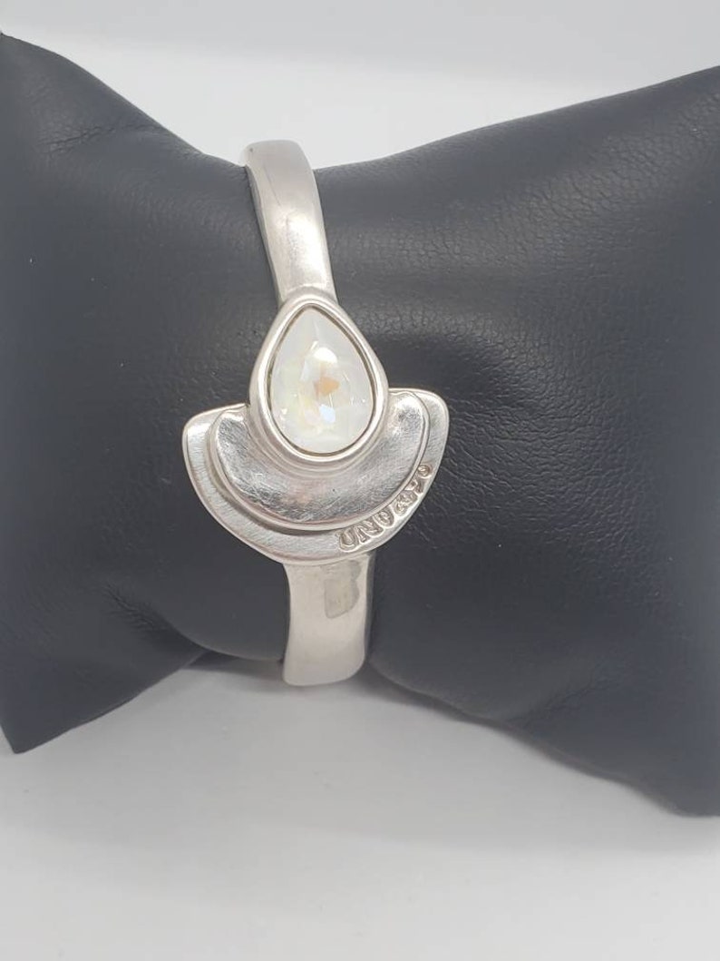 May include: A silver-toned cuff bracelet with a pear-shaped, iridescent crystal centerpiece. The crystal is set within a fan-shaped design. The bracelet is displayed on a black surface.