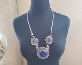 Puede incluir: Collar plateado con tres colgantes circulares y una cadena. Los colgantes presentan piedras grandes, redondas y azul oscuro engastadas en una ornamentación metálica. El collar se exhibe en un maniquí negro.