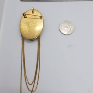 May include: Gold-toned oval brooch with three dangling chain strands. The brooch has a pin closure on the back and a small loop at the top. A coin is next to the brooch for size comparison.