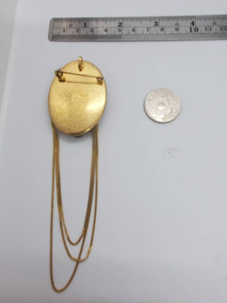 May include: Gold-toned oval brooch with three dangling chain strands. The brooch has a pin closure on the back and a small loop at the top. A coin is next to the brooch for size comparison.