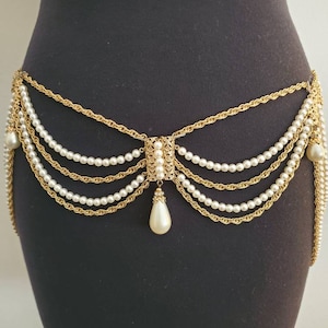 May include: Gold-tone waist belt with multiple strands of faux pearls and chains. The belt features a central decorative element and a teardrop-shaped pearl pendant. The belt is displayed on a black mannequin.