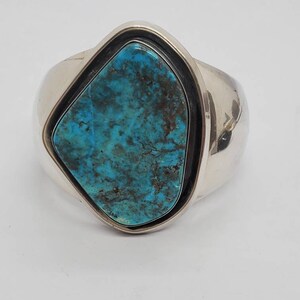 May include: A wide silver cuff bracelet featuring a large, irregular turquoise stone. The stone is a vibrant blue-green color with dark brown matrix patterns. The bracelet has a polished finish and a simple, elegant design.