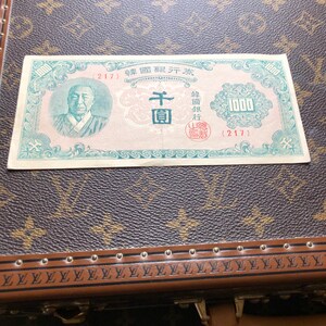 May include: A vintage Korean 1000 Won banknote, featuring a portrait of a man and intricate designs. The bill is primarily light green and pink, with Korean characters and the number 1000 prominently displayed. The banknote is on a brown surface.