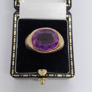 May include: A gold ring with a large, oval-cut purple gemstone, presented in a black jewelry box. The ring's band has a textured design. The box is lined with white satin and has gold trim.