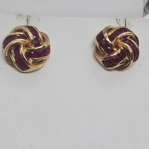 May include: A pair of gold-toned stud earrings. Each earring features a knot design with a series of small, deep red gemstones set within the gold. The earrings are displayed on a white surface.