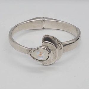 May include: A silver-toned hinged bangle bracelet with a pear-shaped iridescent stone. The bracelet has a curved design with a decorative element featuring the stone and a curved accent. The word "ORIENT" is engraved on the accent.