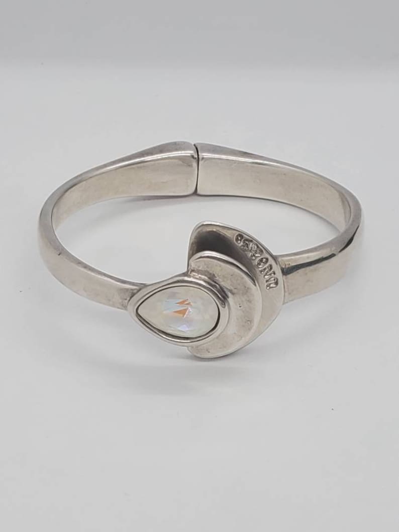 May include: A silver-toned hinged bangle bracelet with a pear-shaped iridescent stone. The bracelet has a curved design with a decorative element featuring the stone and a curved accent. The word "ORIENT" is engraved on the accent.